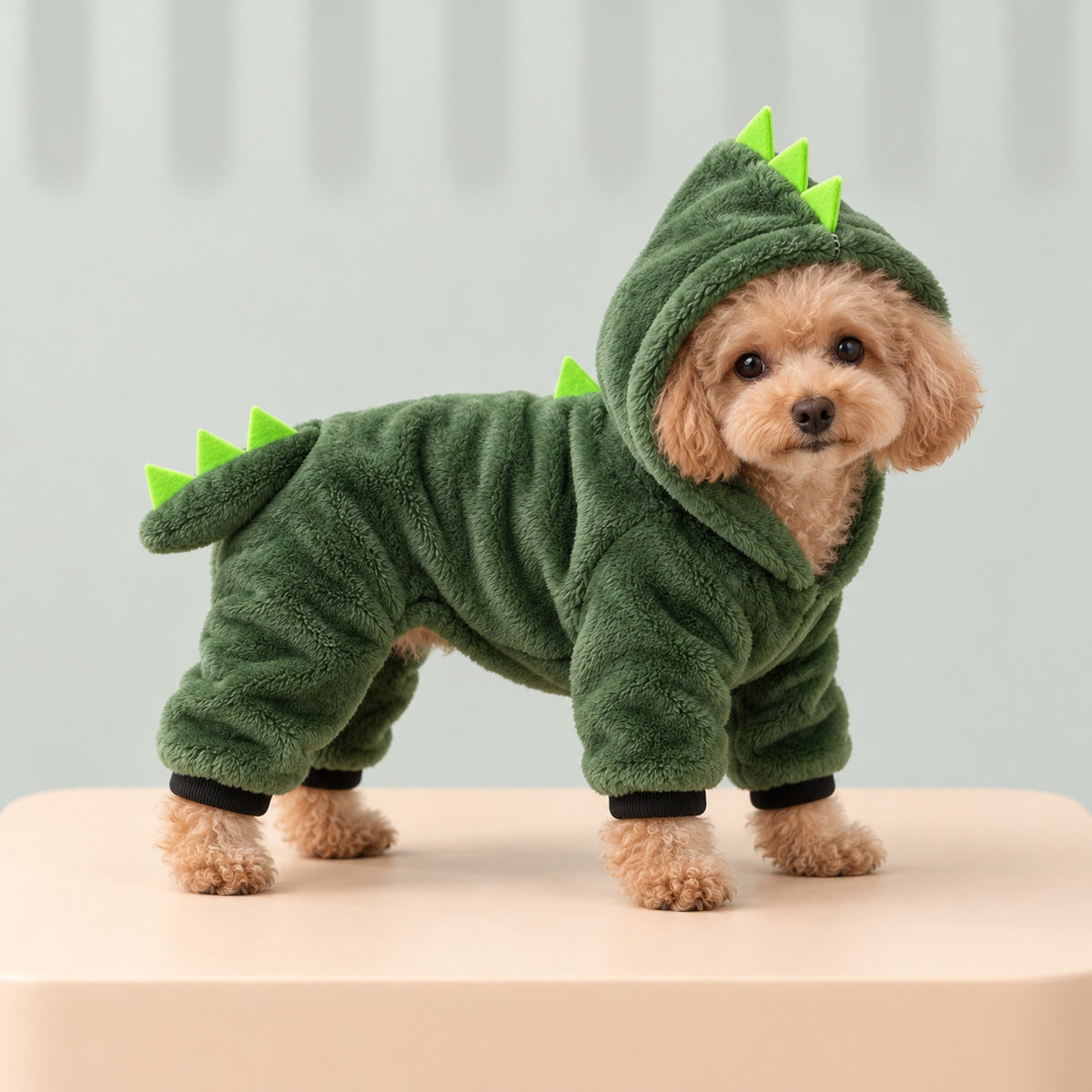 Apricot toy poodle wearing the green dinosaur jumpsuit with the spiked hood up